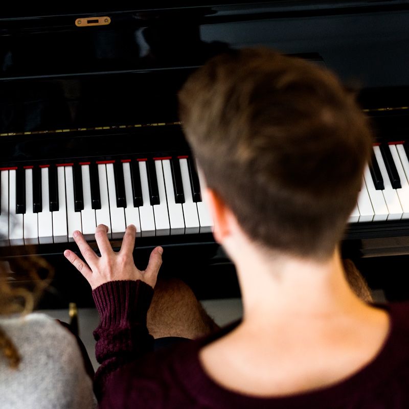 An adult student playing an upright piano, seen from over the shoulder
